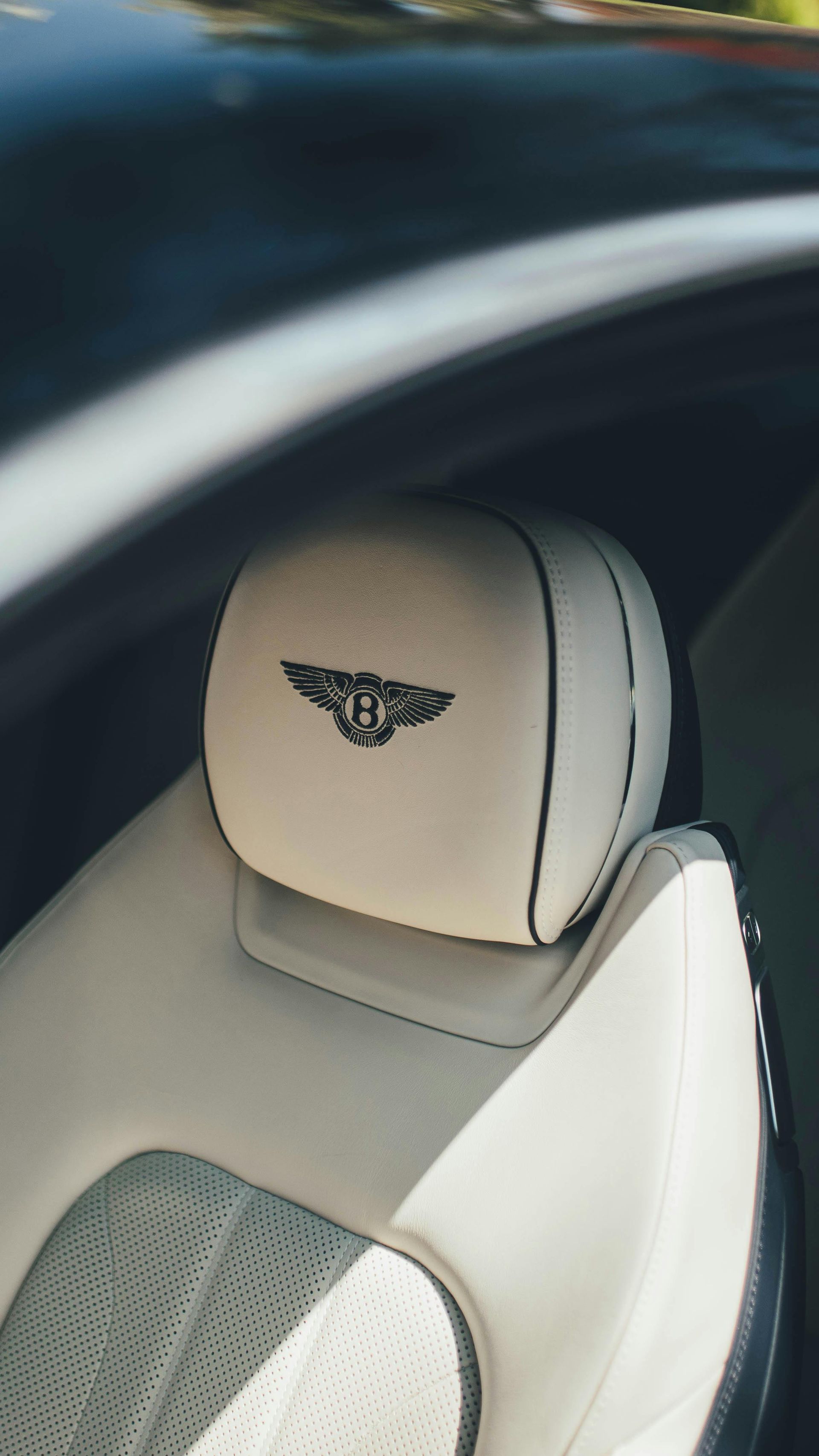 A cream-colored leather car seat headrest with an embroidered Bentley logo in a dark, luxury vehicle interior.