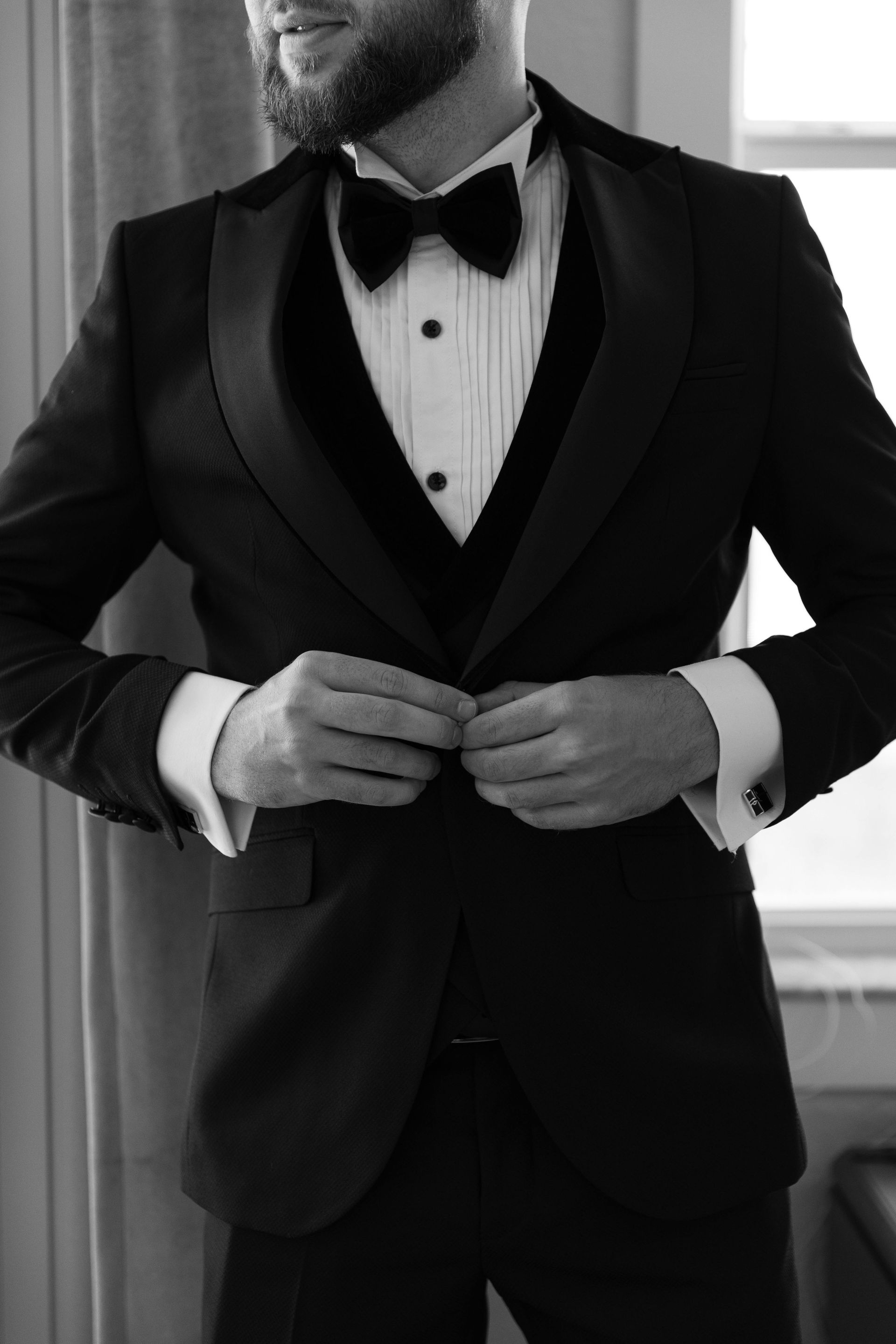 A bearded person adjusting the button of a black tuxedo, complete with a bowtie and white pleated dress shirt.
