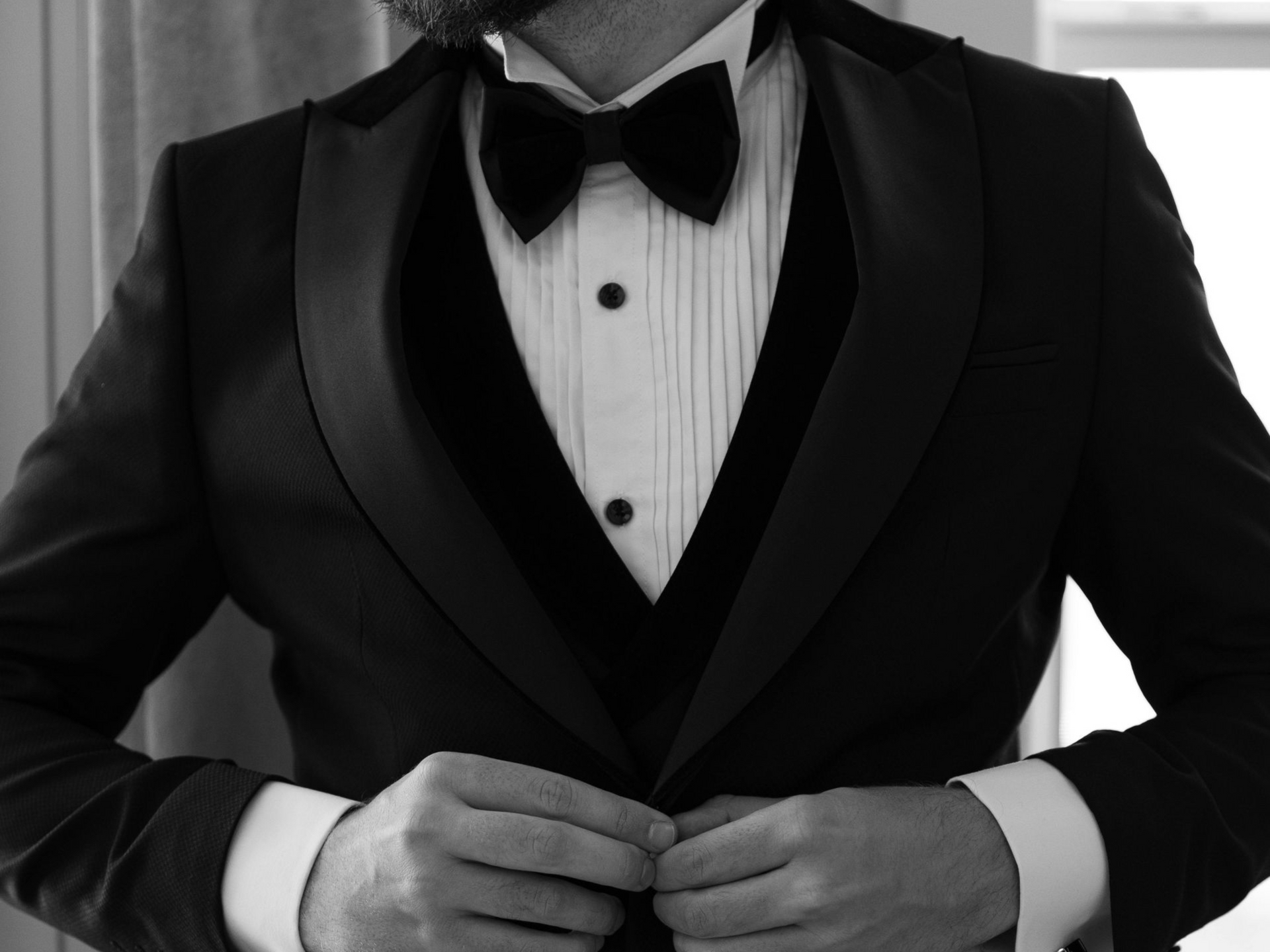 A person in a black tuxedo with a bowtie, adjusting the buttons on their vest.