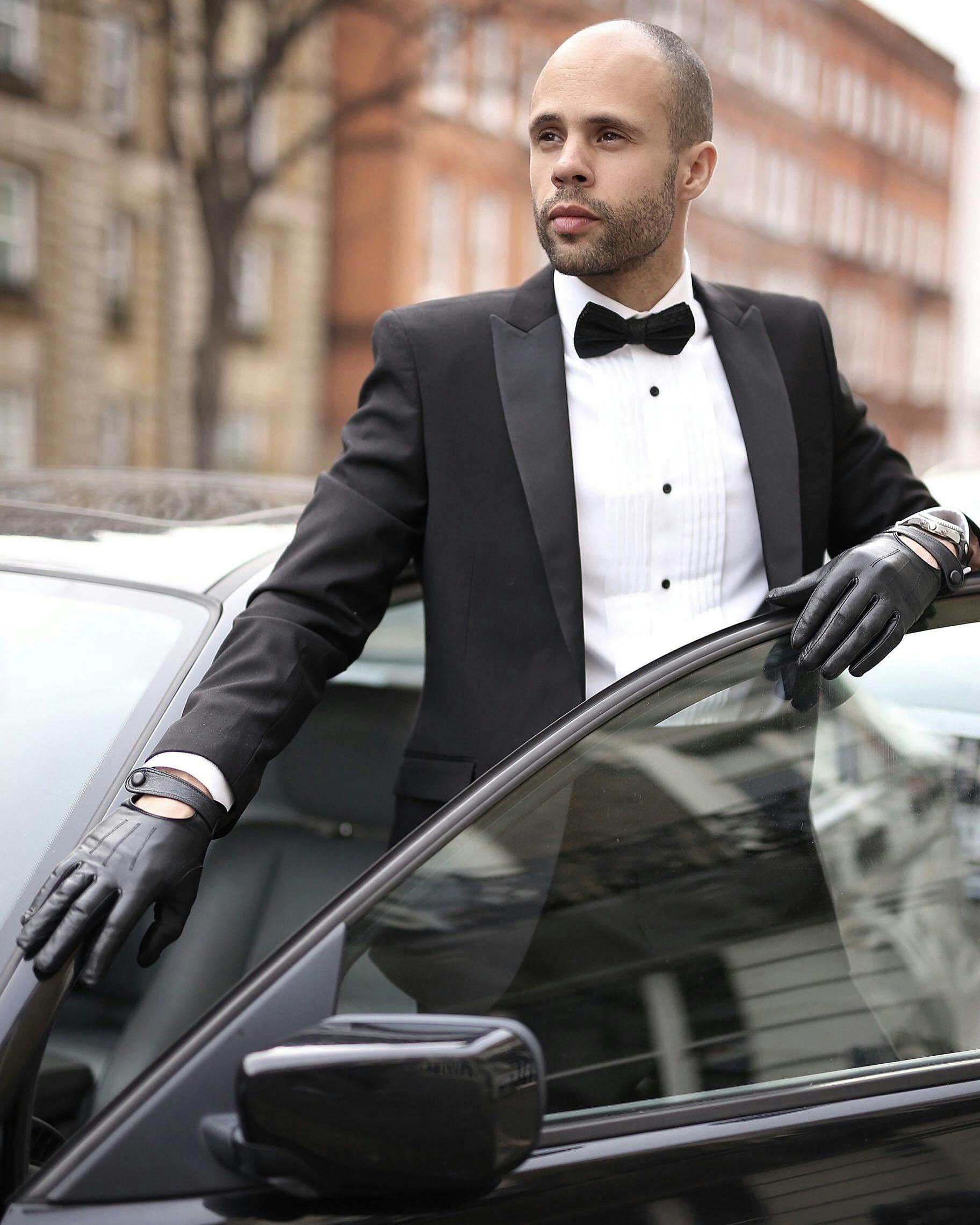 A person in a black tuxedo and bowtie with leather driving gloves leaning against a car door on a city street.