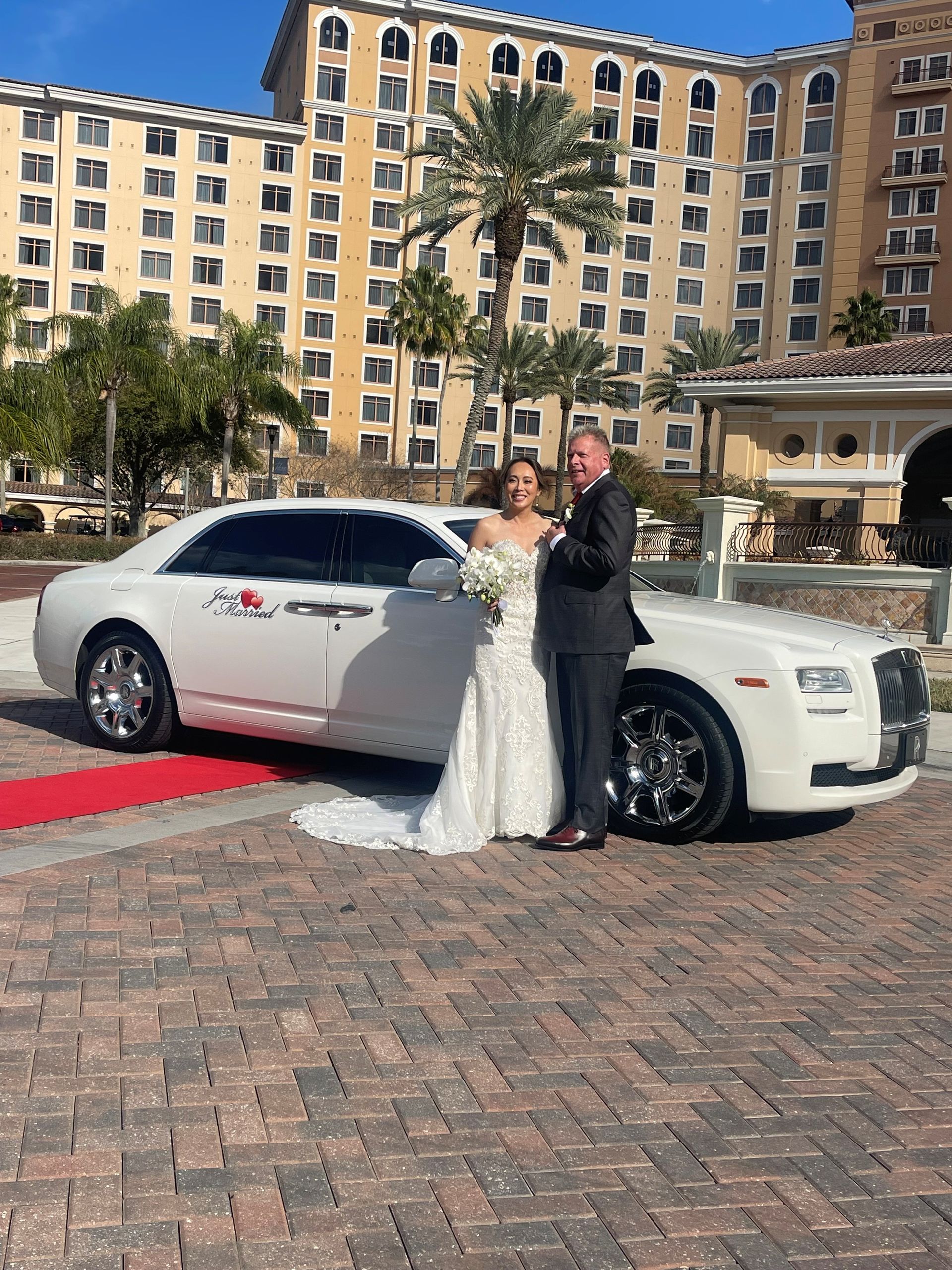 A couple in wedding attire stands on a red carpet next to a long, white Rolls-Royce outside a large resort hotel.