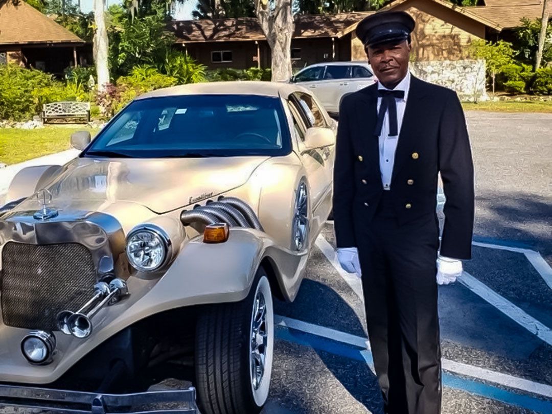 A chauffeur in a black tuxedo and cap stands beside a vintage-style tan car in an outdoor parking lot.