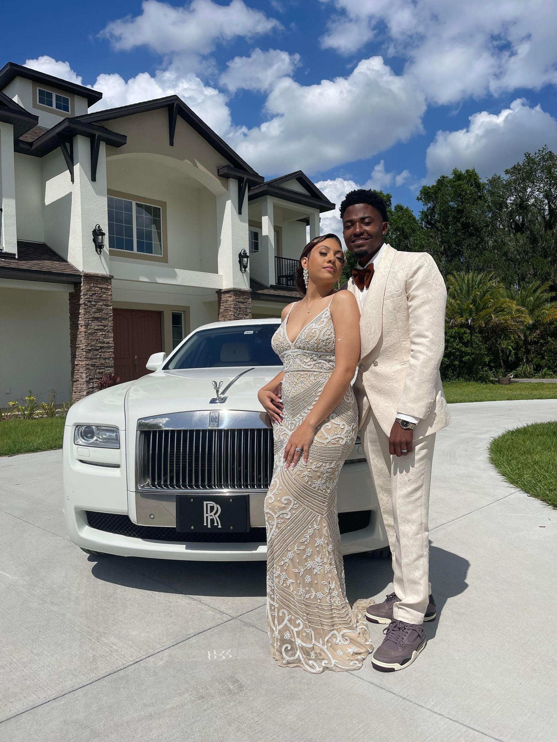 A couple in formal attire stands in front of a white luxury car parked outside a multi-story house on a sunny day.