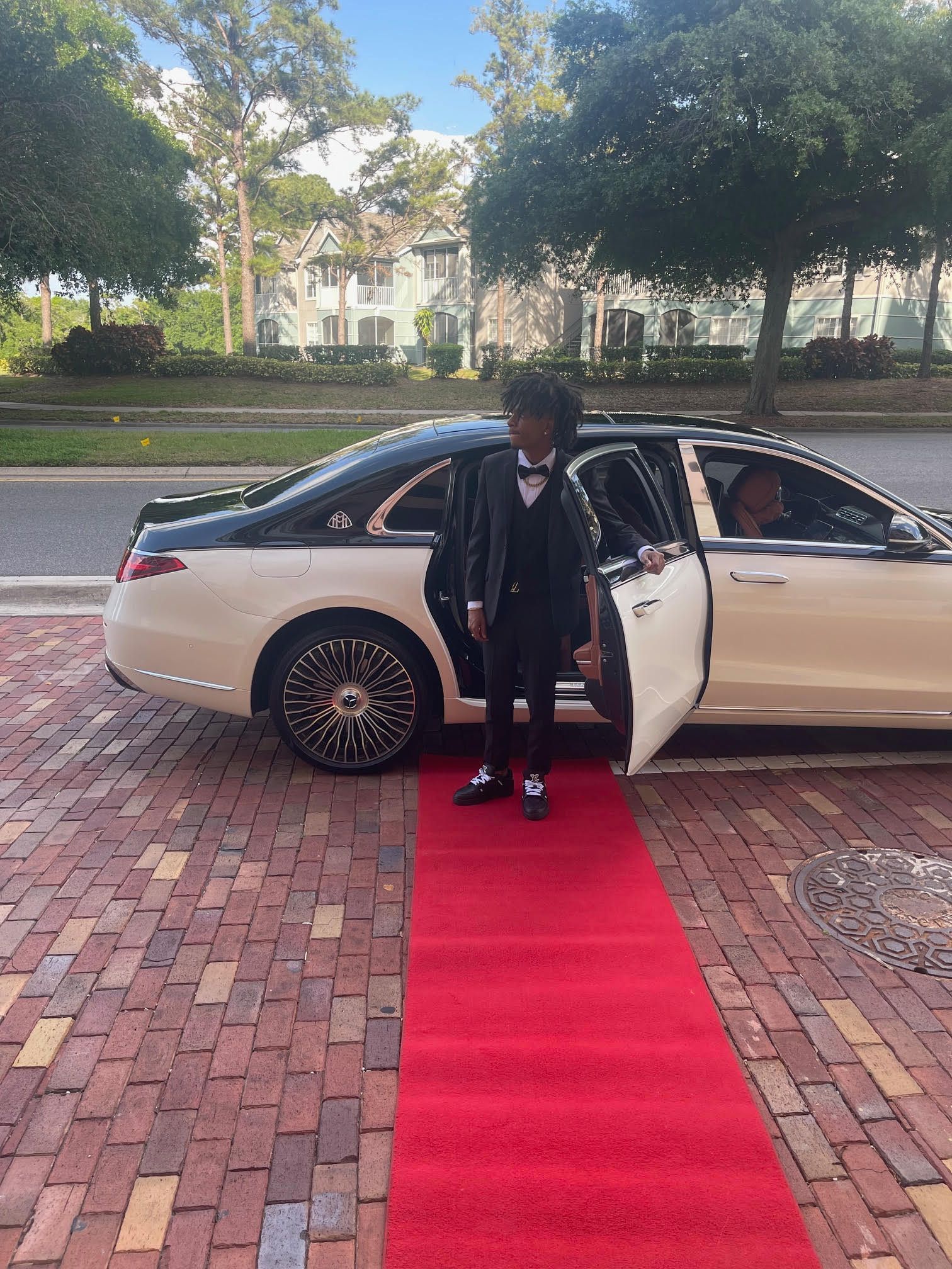 A person in a formal suit stands on a red carpet next to an open door of a two-tone, white and black luxury sedan.