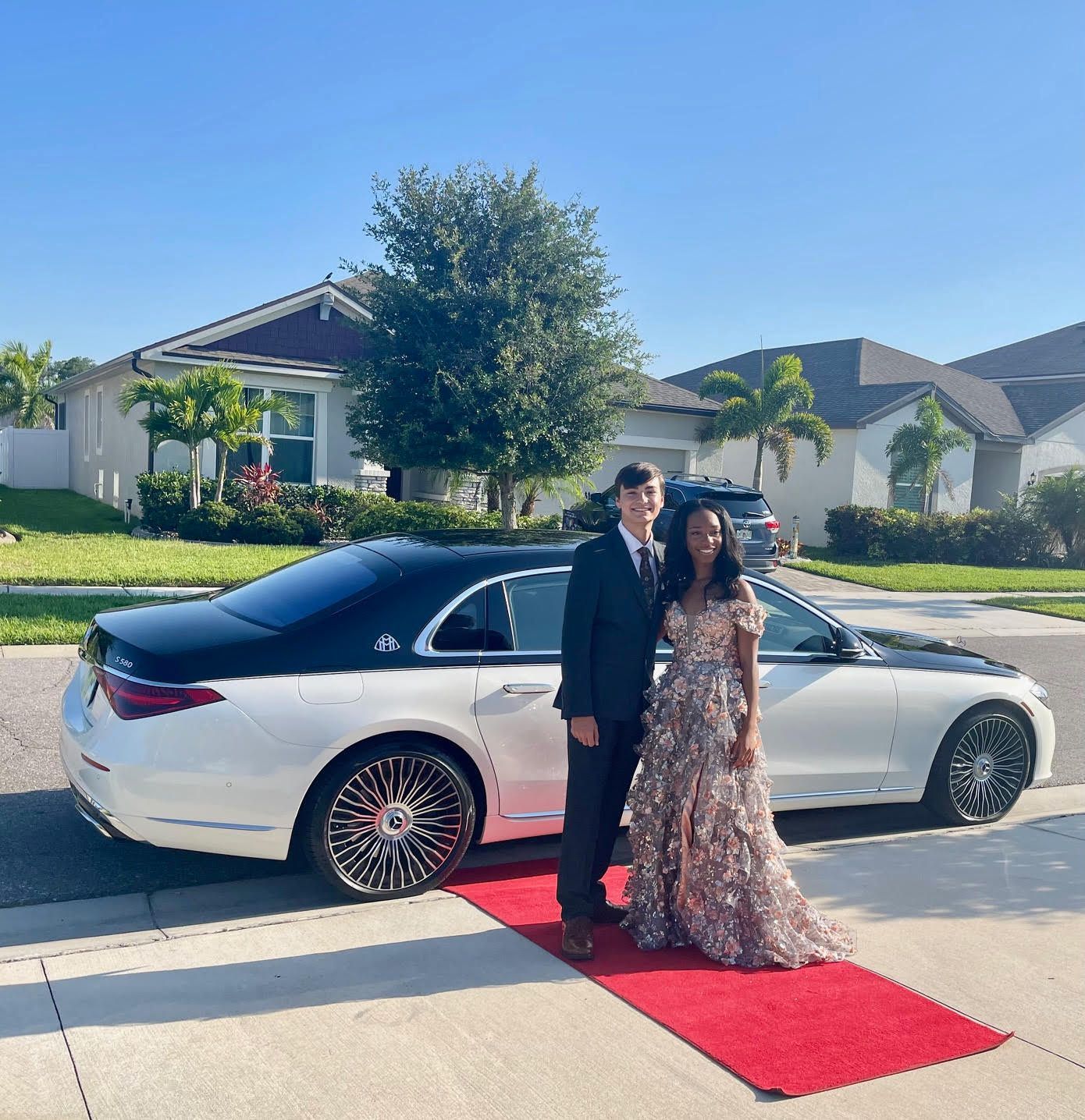 A couple in formal attire stands on a red carpet in front of a white and black luxury sedan in a residential driveway.