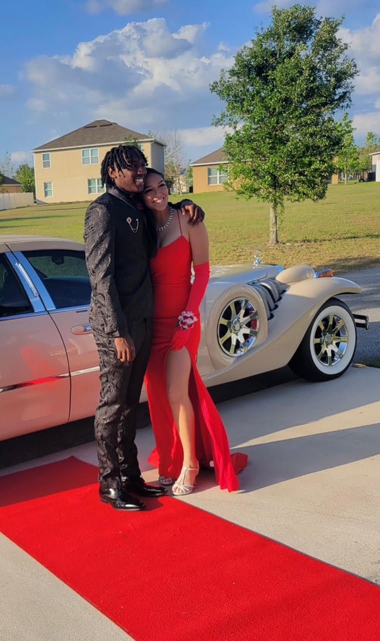 A couple stands together on a red carpet next to a vintage cream-colored car outside on a sunny day.