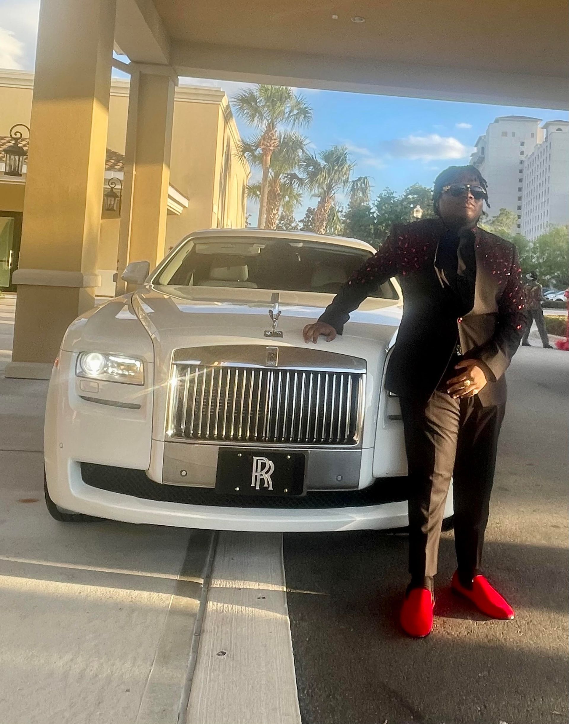 A person in a dark patterned suit and red shoes stands next to a white Rolls-Royce parked under a building overhang.