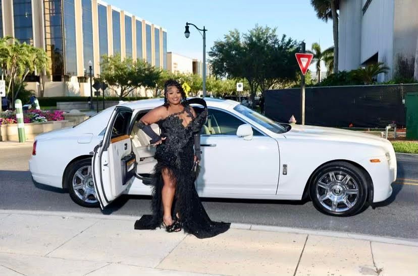 A person in a black, feathered, high-slit gown stands beside an open door of a white luxury car on a sidewalk.