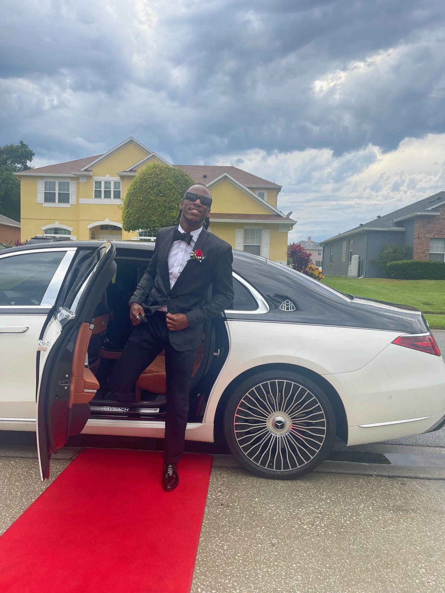 A person in a black tuxedo and sunglasses steps out of a white luxury car onto a red carpet in front of a suburban home.