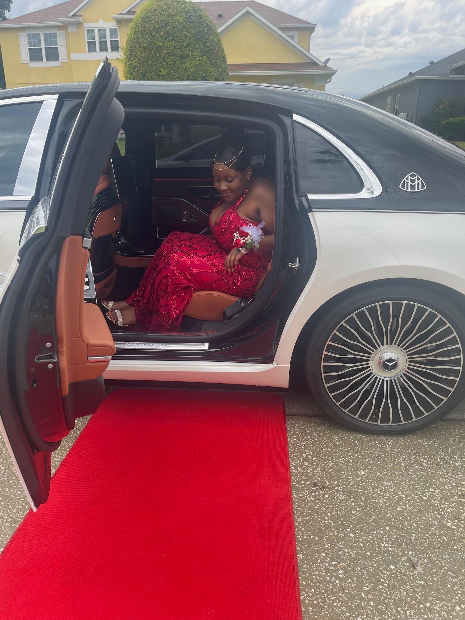 A person in a red sequined gown steps onto a red carpet from the open door of a white luxury sedan.