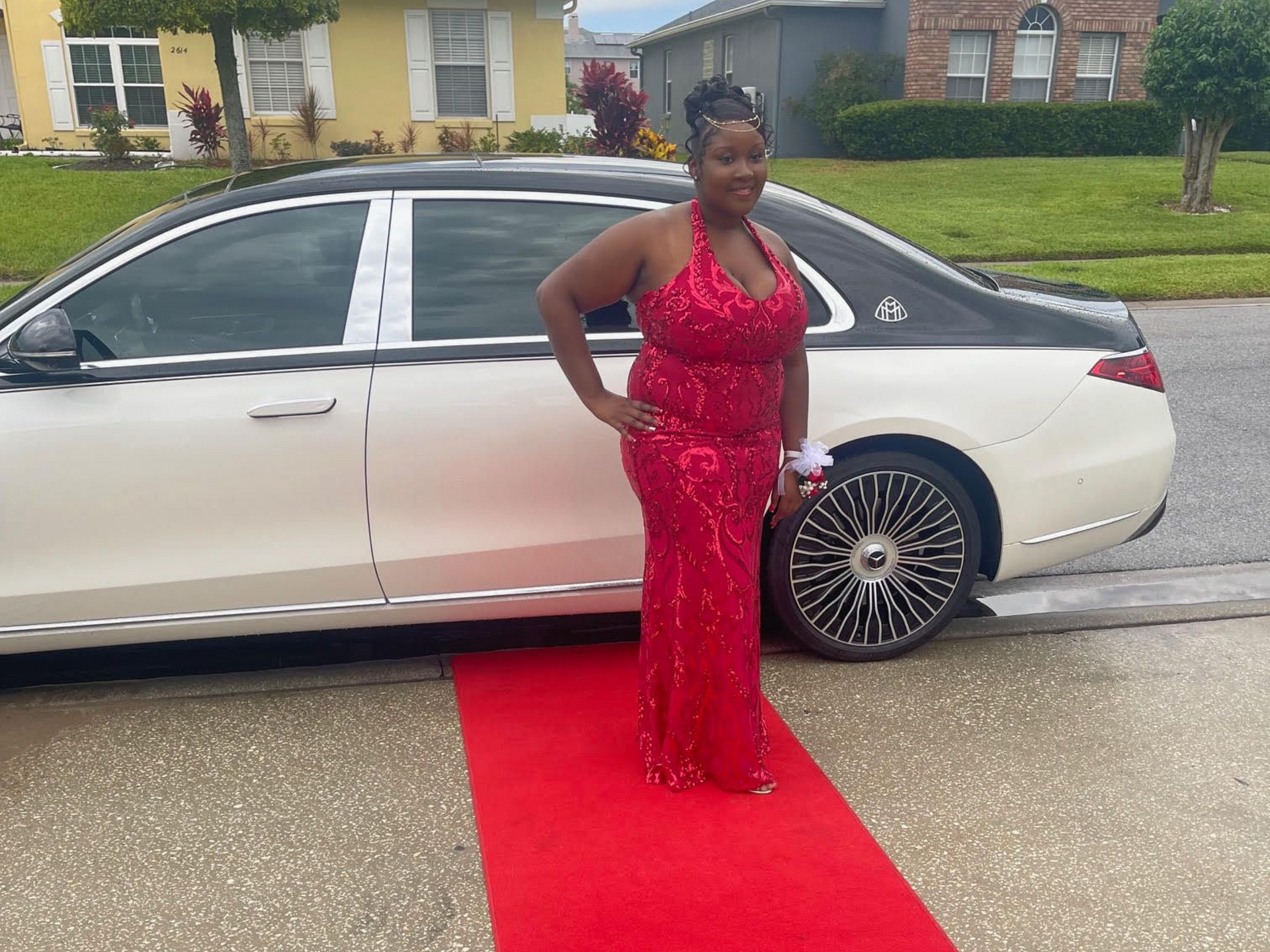 A person in a red sequined gown stands on a red carpet in front of a white and black luxury car parked on a street.