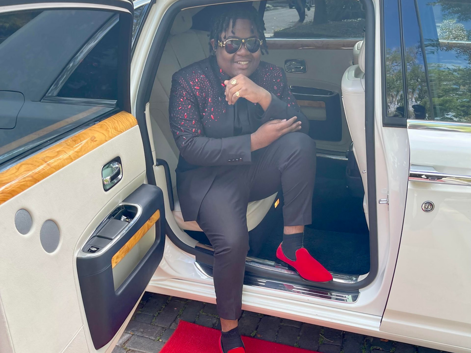 A person sits in an open luxury car door, wearing a patterned dark blazer, dark trousers, and bright red loafers.