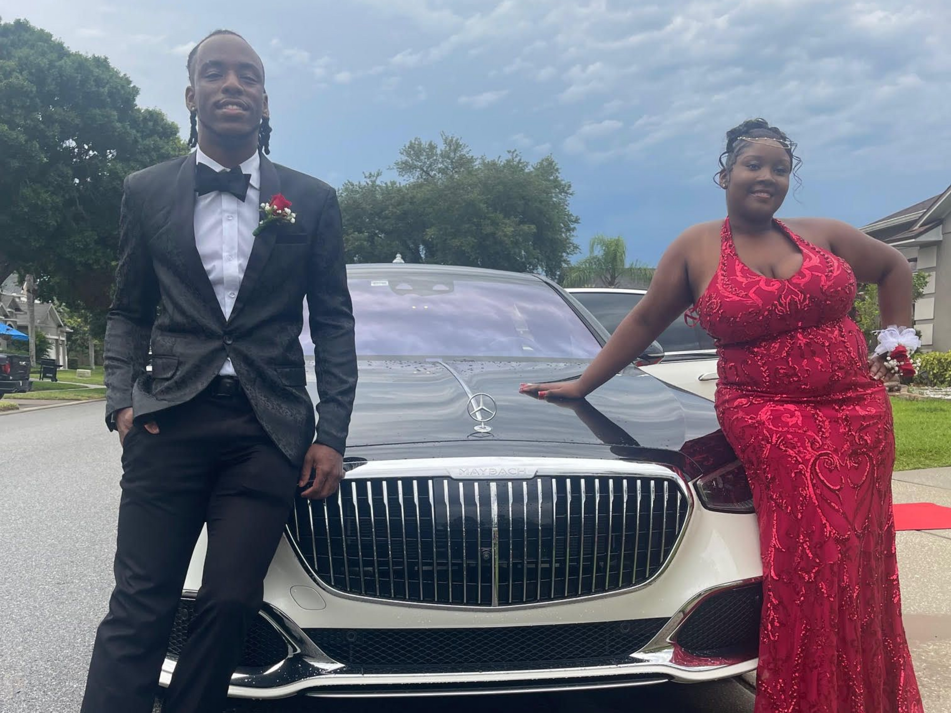 A person in a black tuxedo and a person in a red sequined gown pose smiling together in front of a luxury car.