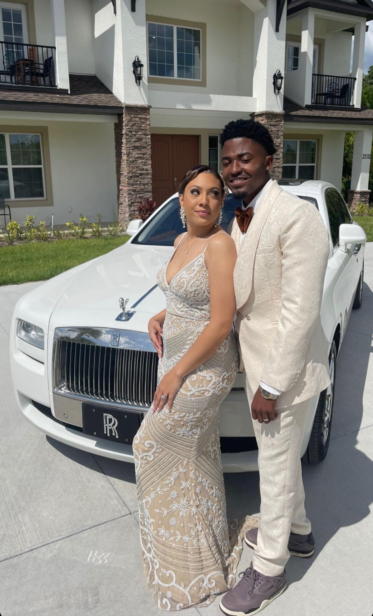 A couple in elegant formal attire poses together in front of a white luxury car and a large, multi-story house.