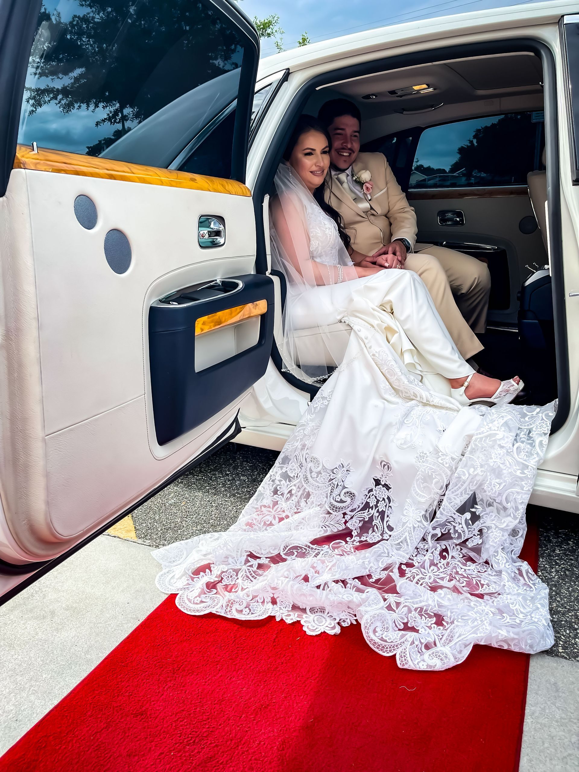 A couple in formal wedding attire seated inside a white limousine, the bride's long lace train draped onto a red carpet.
