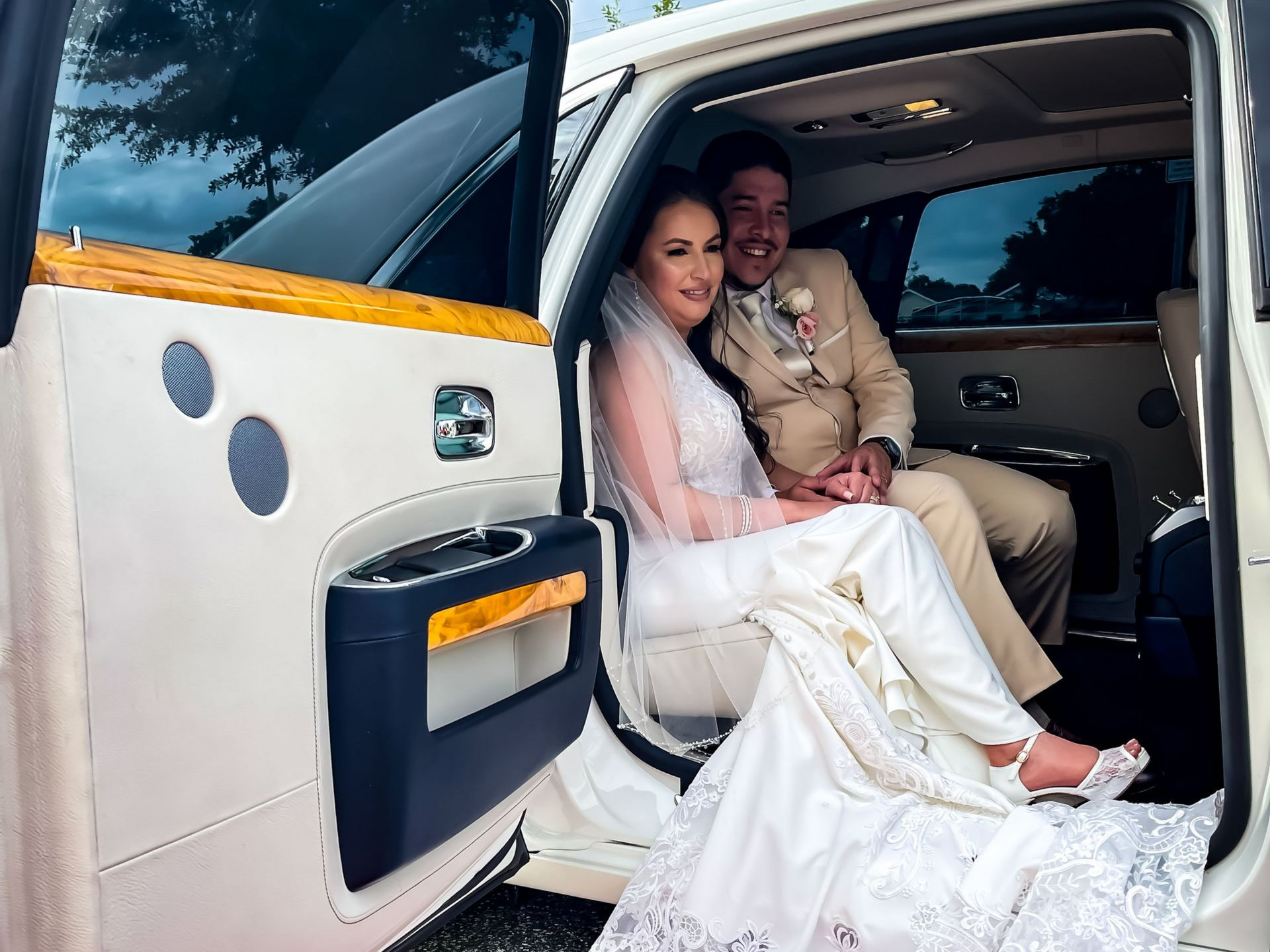 A bride and groom sitting inside a luxury car with the door open, looking towards the camera and smiling.