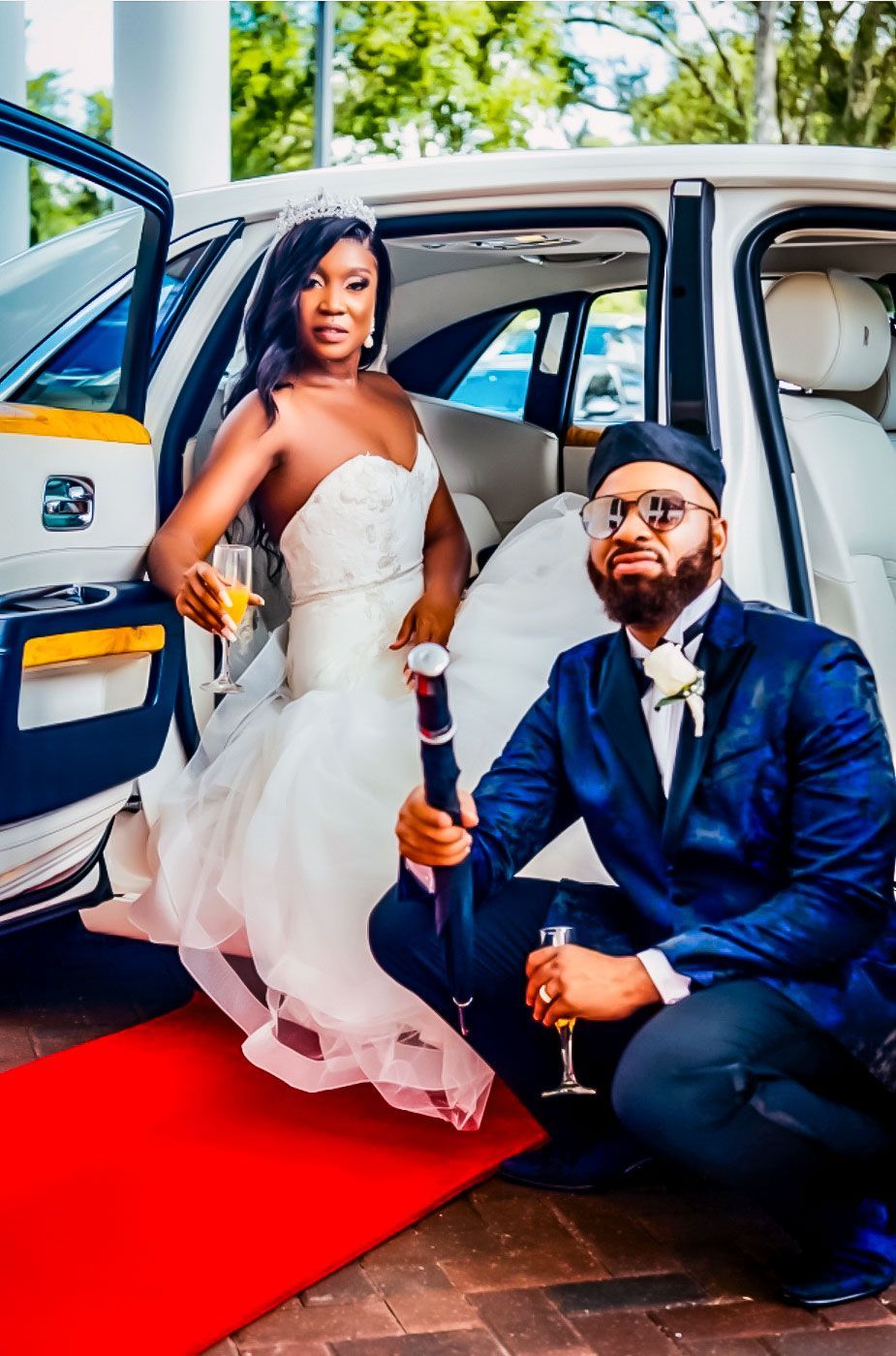 A bride stands by a white car as a person in a blue suit kneels on a red carpet, holding a party popper and glass.