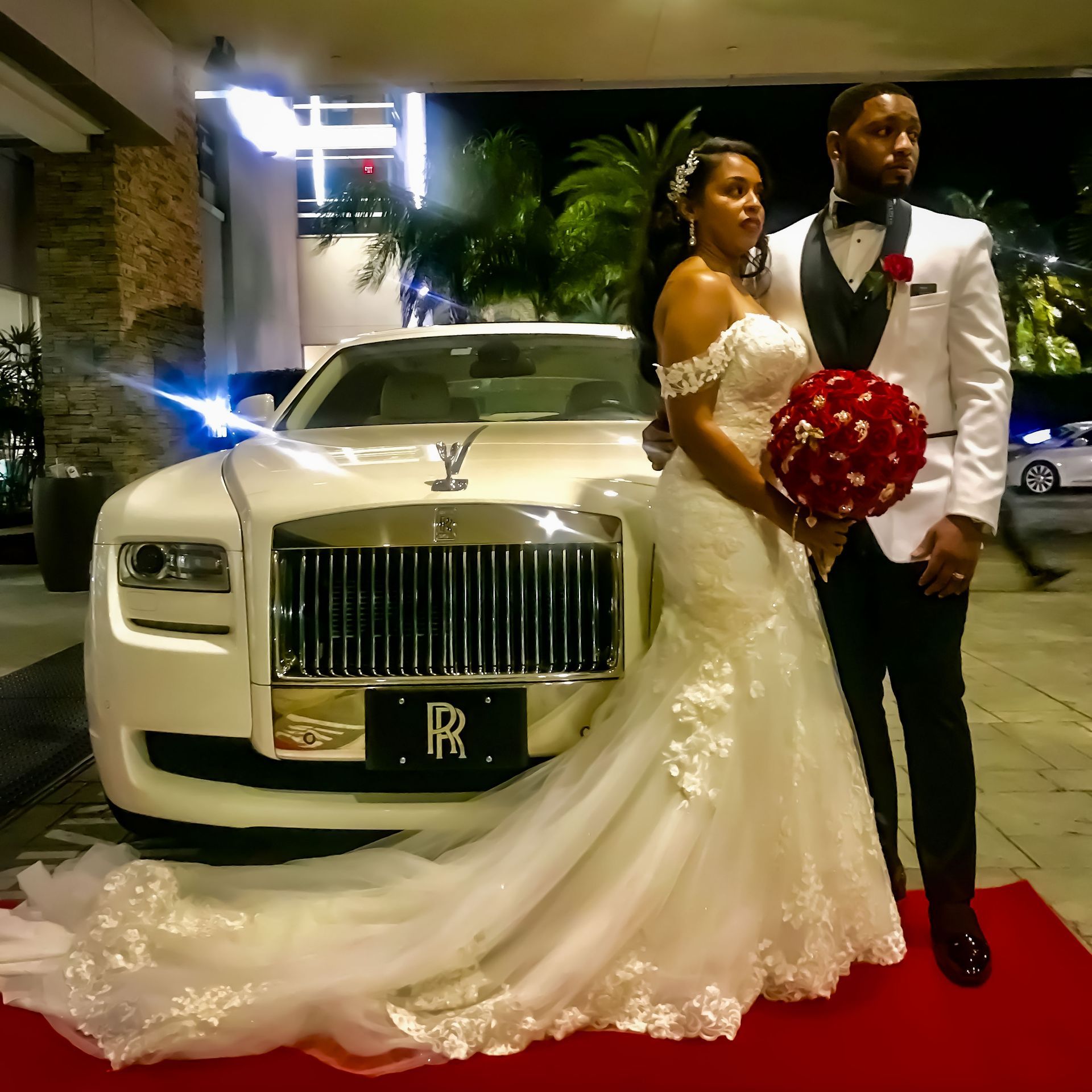 A bride and groom stand on a red carpet next to a white Rolls-Royce at night.