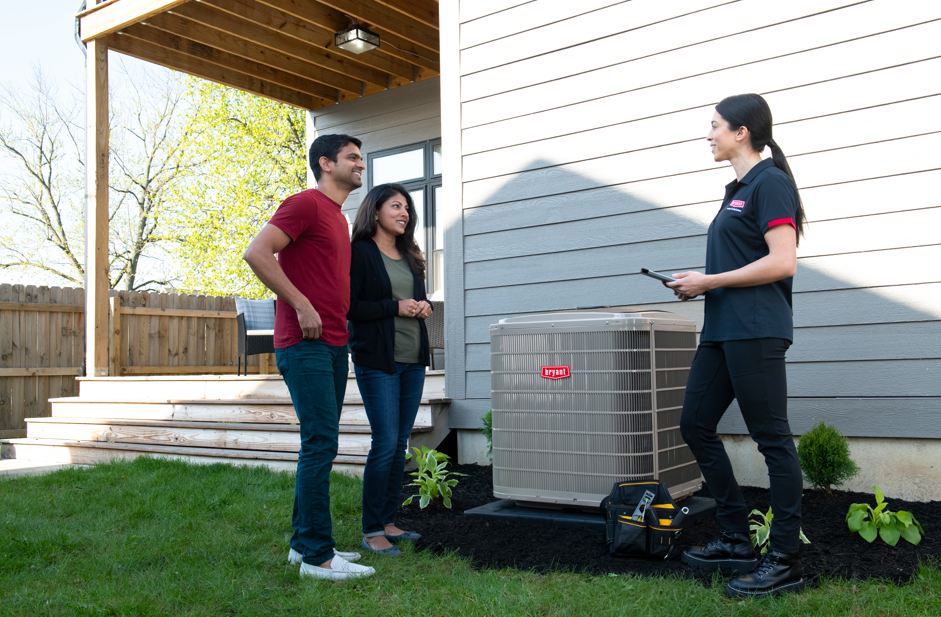 Three people stand beside an outdoor air conditioner unit next to a house.