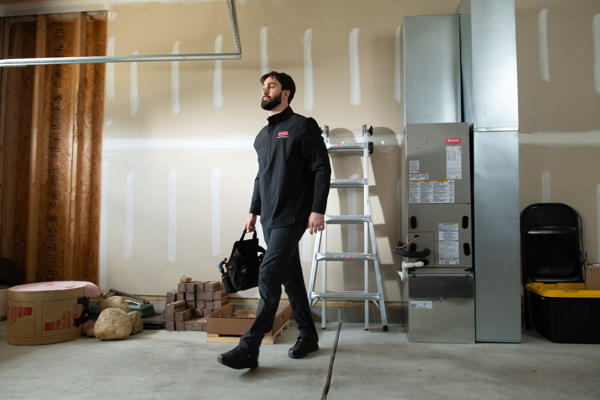 Person carrying a tool bag in a garage beside a ladder and electrical panel