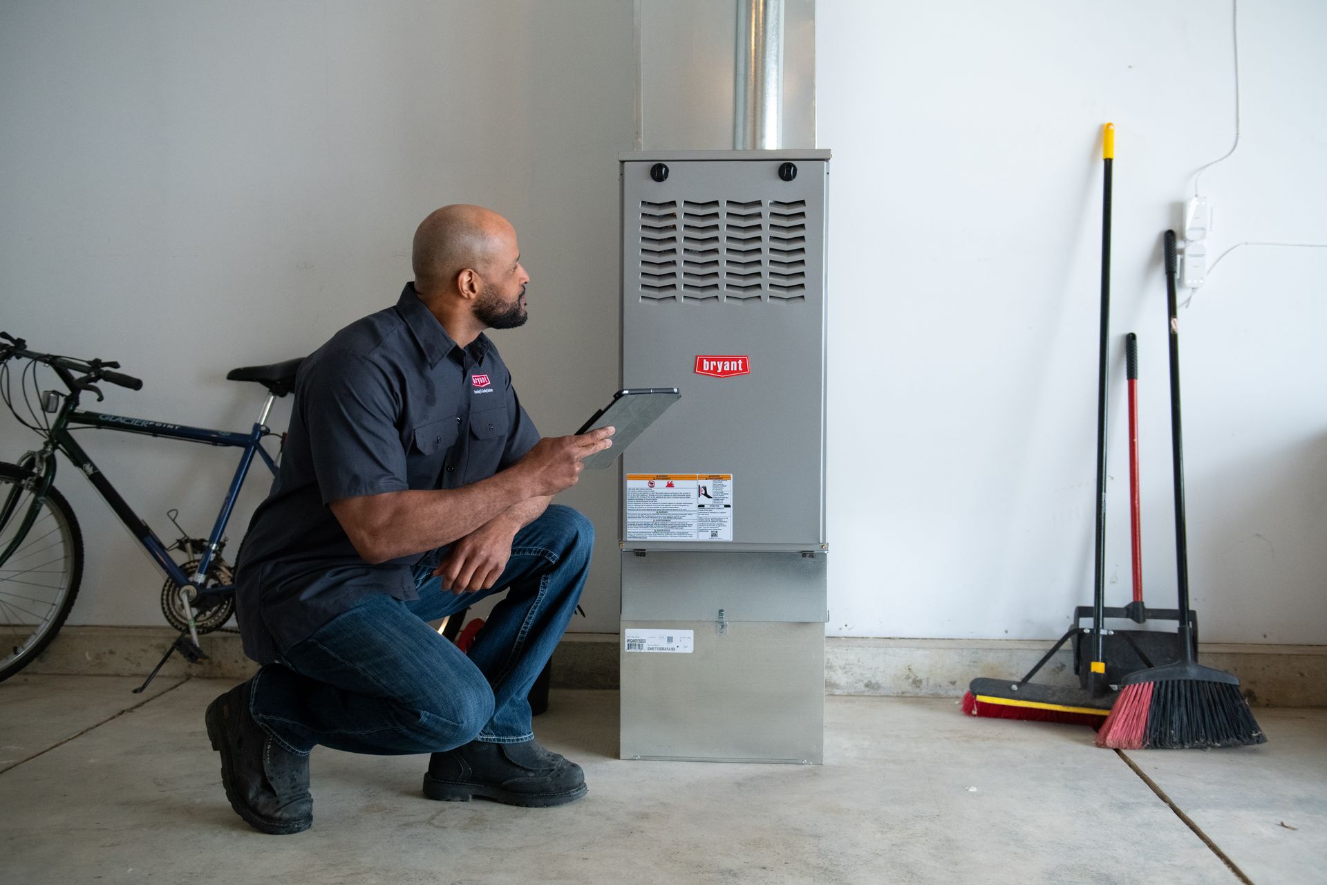 Man kneeling beside a furnace in a garage, inspecting it with a clipboard; bicycle and cleaning tools nearby.