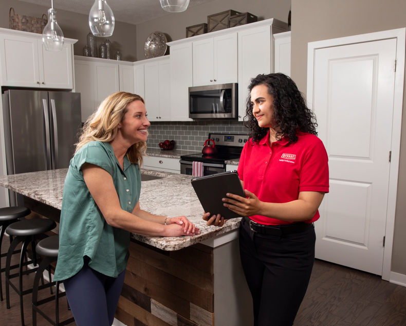 Two women talk in a modern kitchen while one holds a clipboard.