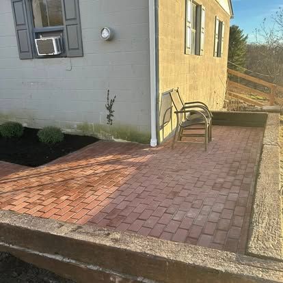 Brick patio beside a beige house with a chair, small garden beds, and a concrete edge in front.