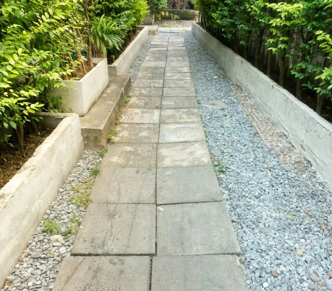 Stone walkway beside a gravel path, bordered by lush green plants on both sides.