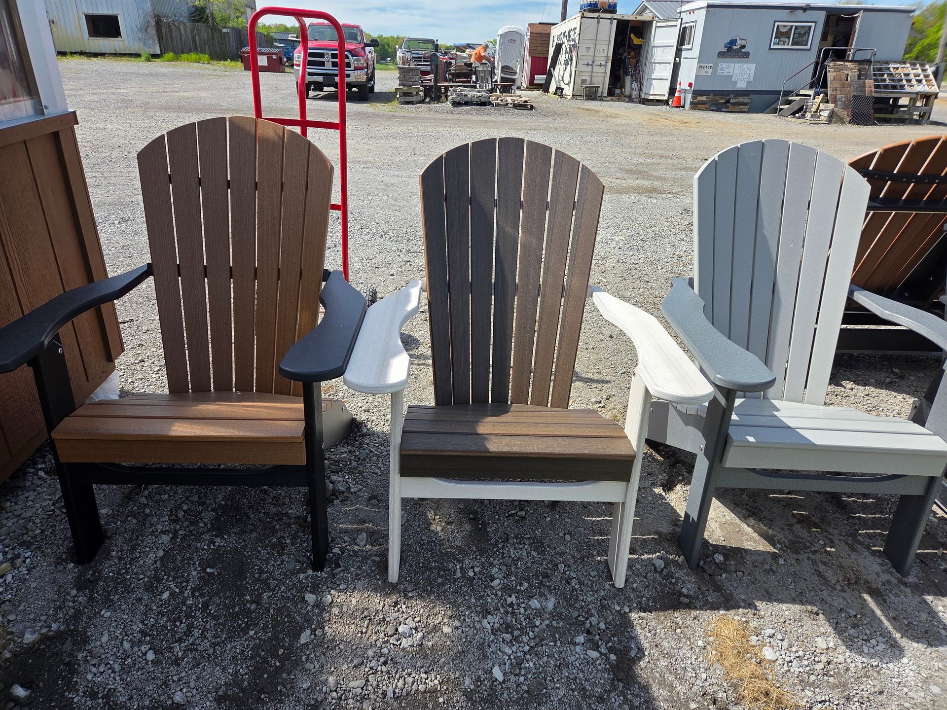 Three colorful Adirondack chairs on gravel outdoors, with trucks and a red hand truck in the background