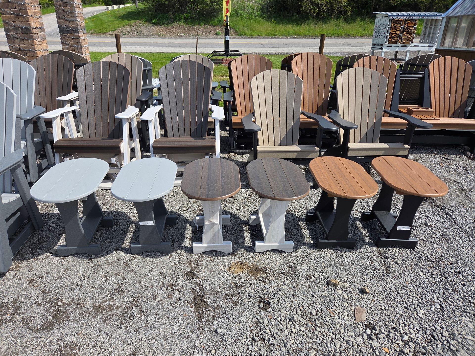 Outdoor display of assorted Adirondack chairs in white, gray, tan, and black on gravel near a road