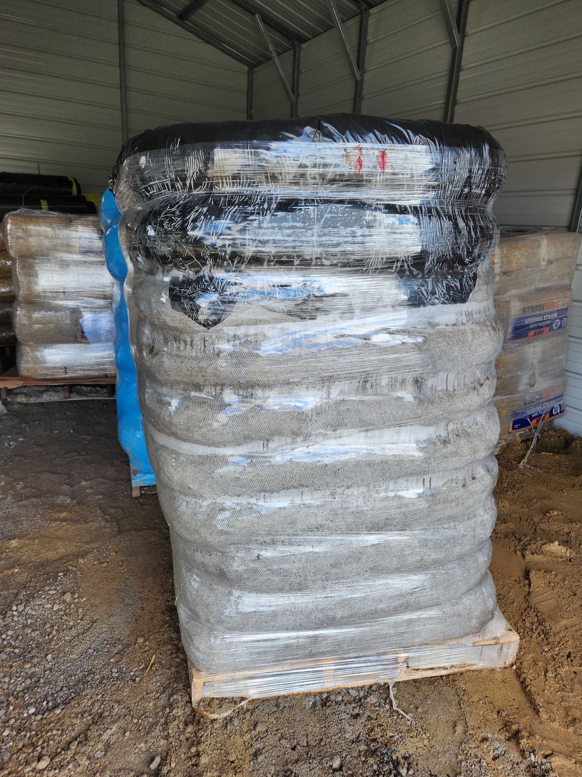 Stack of shrink-wrapped black containers in a warehouse, with other wrapped pallets in the background.