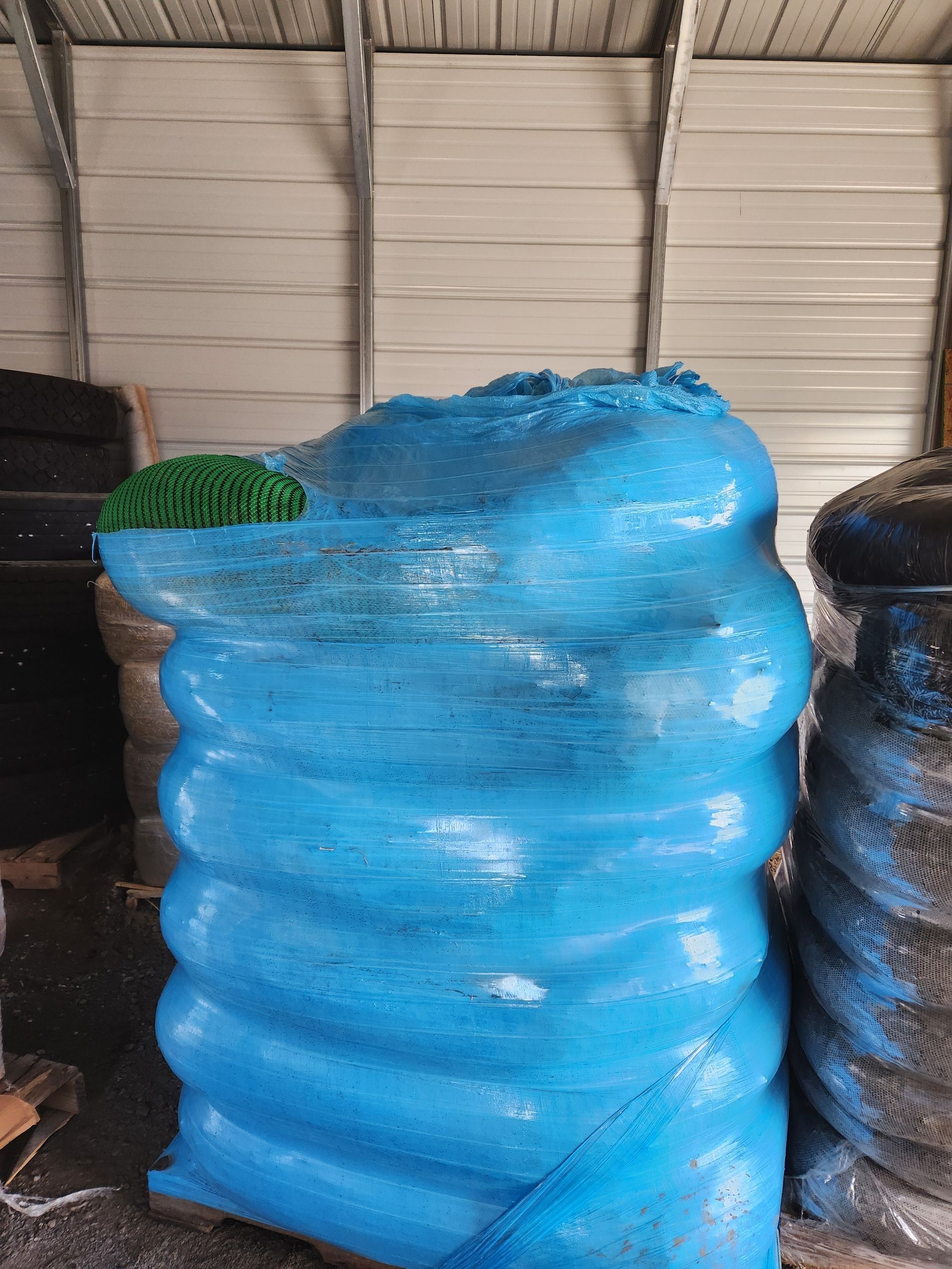 Blue plastic-wrapped stack of bales in a warehouse, with green material visible on top.