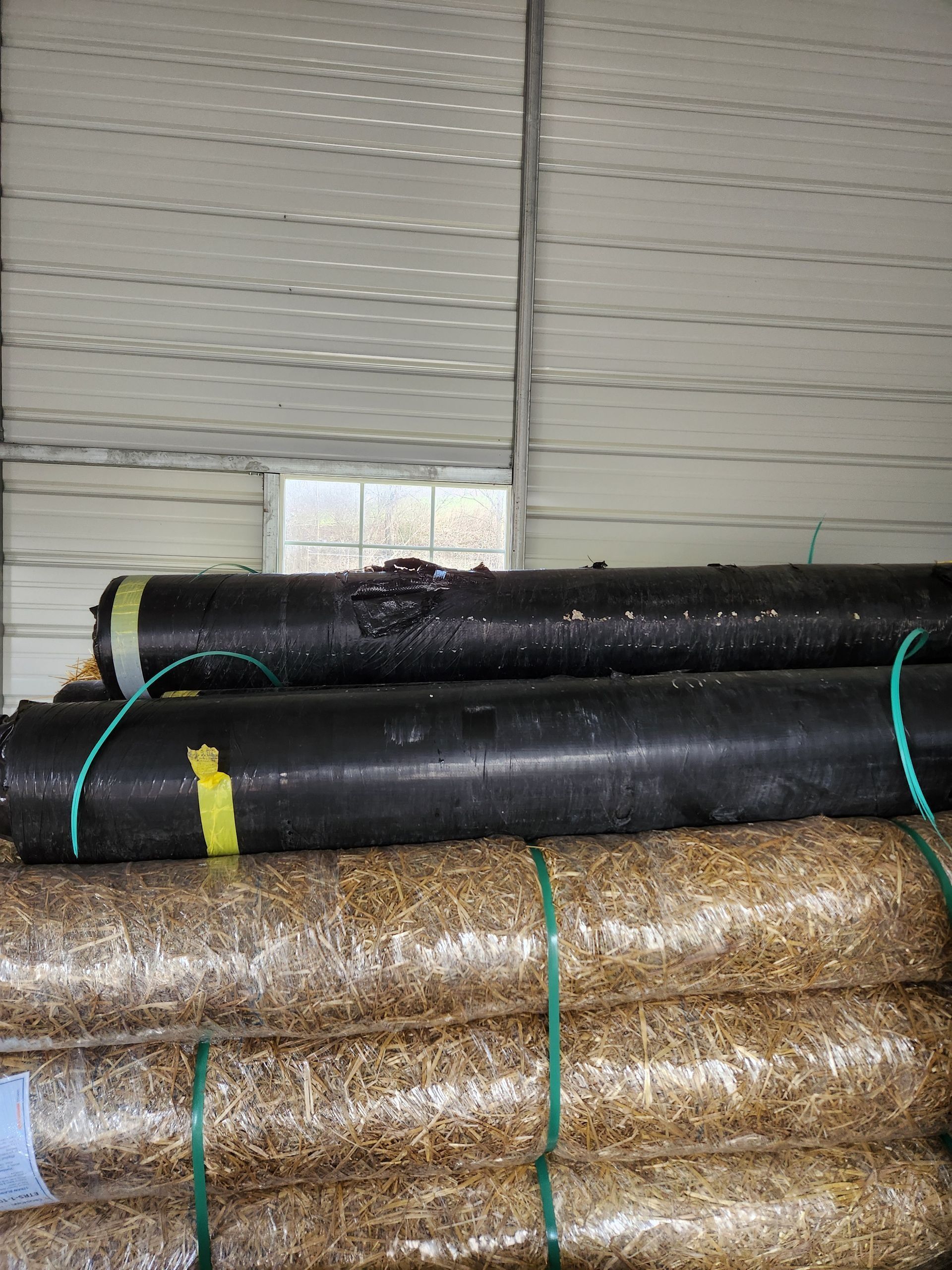 Stacked rolls of black and tan insulation or flooring material in a warehouse against a corrugated metal wall