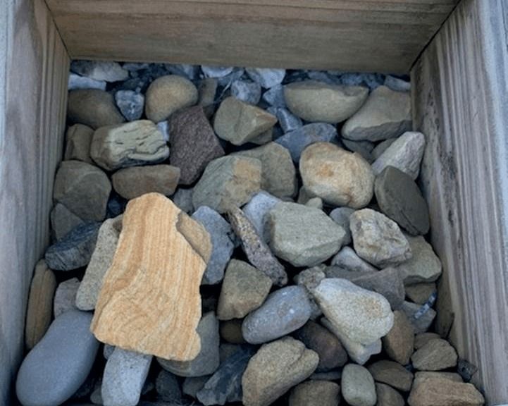 Wooden crate filled with assorted gray rocks, with one tan rock in front.