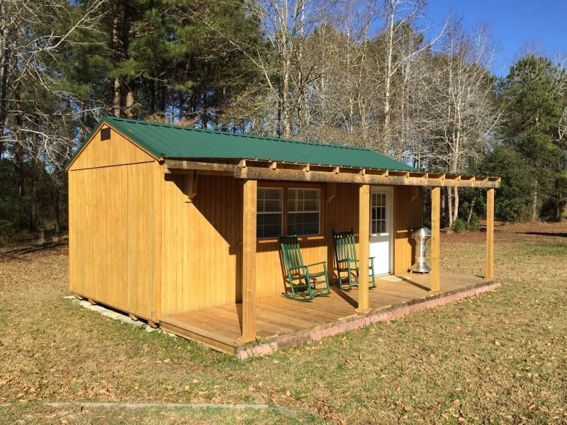 Small wooden cabin with green roof and porch chairs in a grassy yard among trees