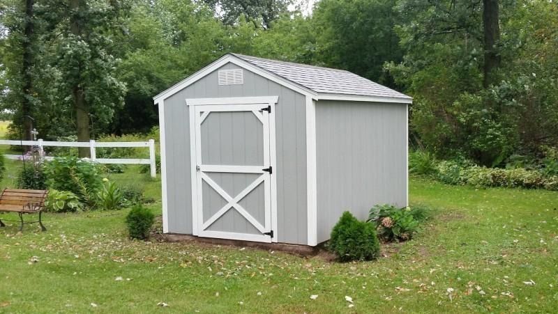 Small gray garden shed with white trim in a grassy yard, surrounded by trees and a white fence.