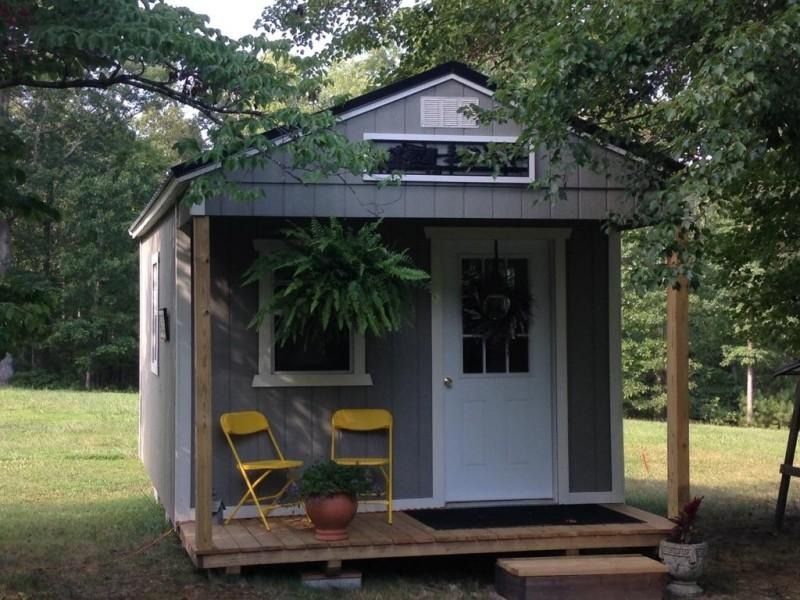 Small gray cottage with white trim, porch, and two yellow chairs under trees.
