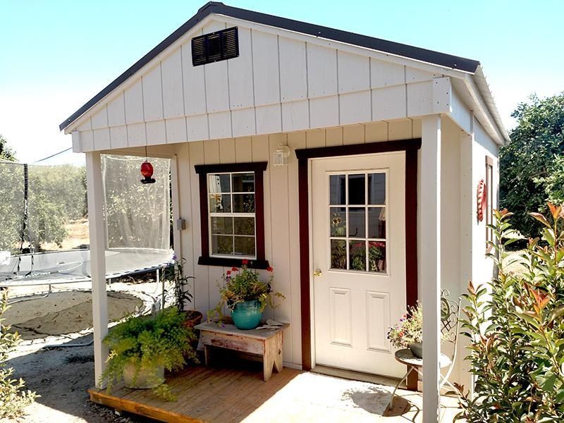 Small white garden shed with porch, black trim, and potted plants beside the door