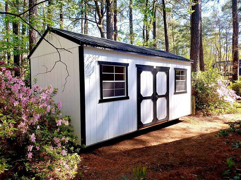 White garden shed with dark roof and doors in a wooded yard, surrounded by pink flowers and trees