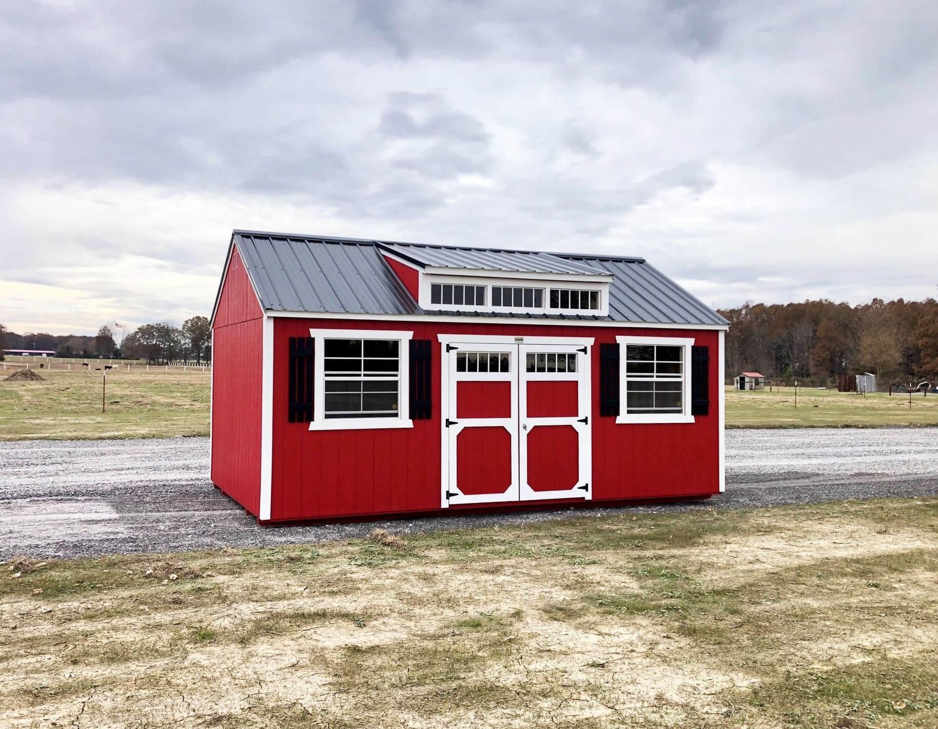 Small red barn-style shed with white trim on a gravel lot under a cloudy sky