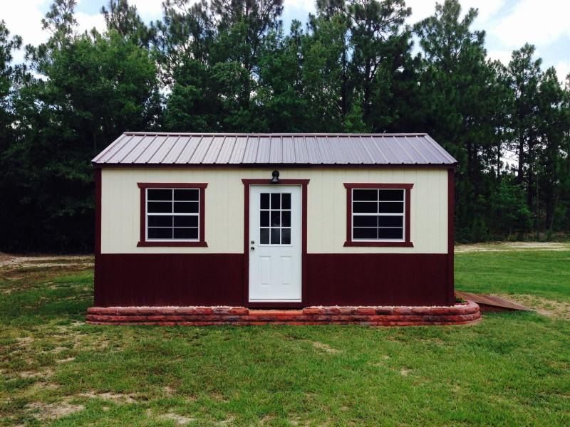 Small cream-and-maroon shed with a white door and windows, set on grass in front of trees.