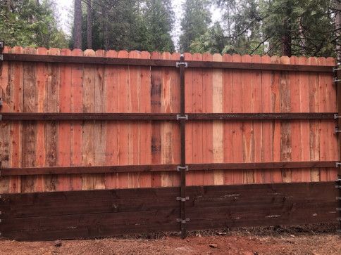 Tall wooden fence with reddish planks and dark brown base, set against trees in the background