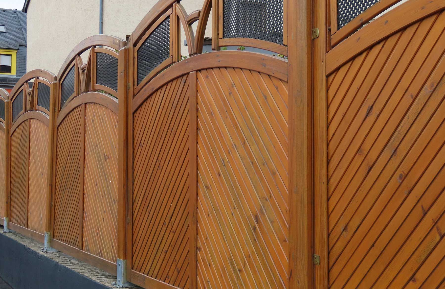 Wooden fence with curved panels along a sidewalk, in warm brown tones.