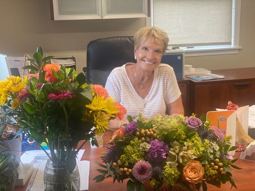 Woman with short blonde hair smiles behind a desk, flanked by two bouquets of colorful flowers.