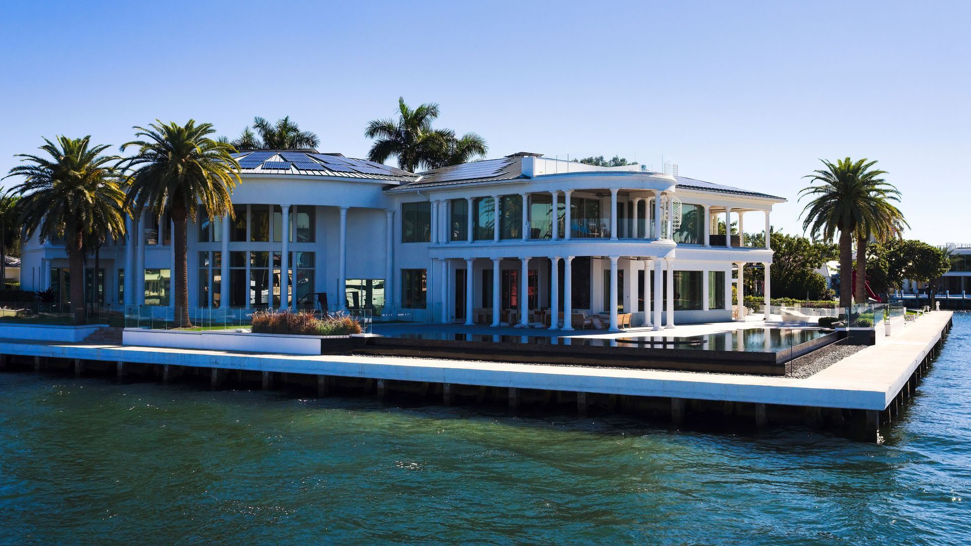 Modern waterfront mansion with palm trees and a dock, beside blue water under a clear sky