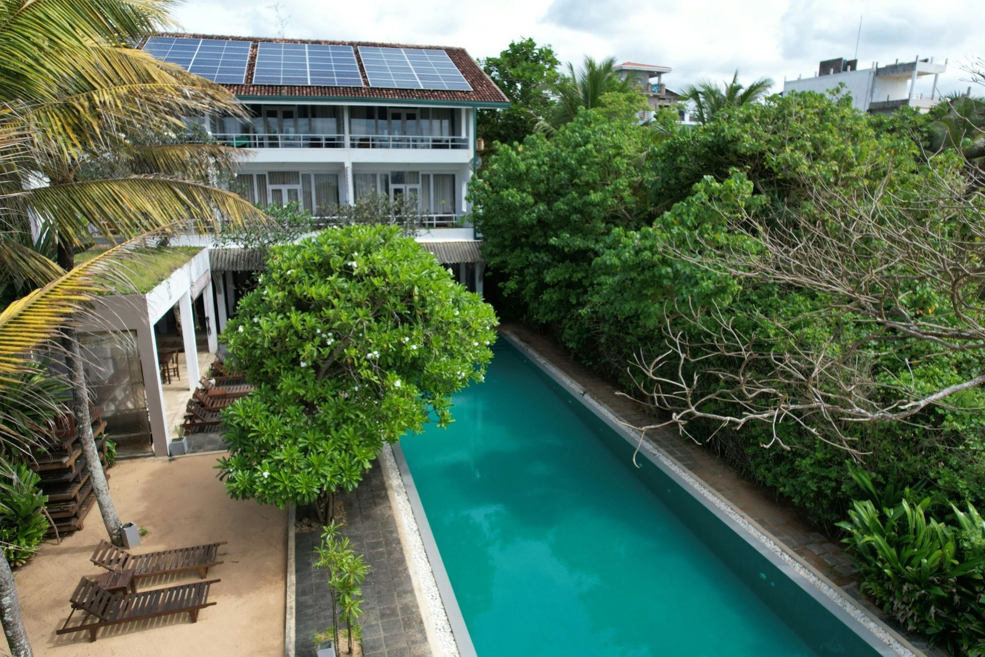 Tropical resort courtyard with a turquoise pool, lush trees, and solar-panelled building overlooking the water