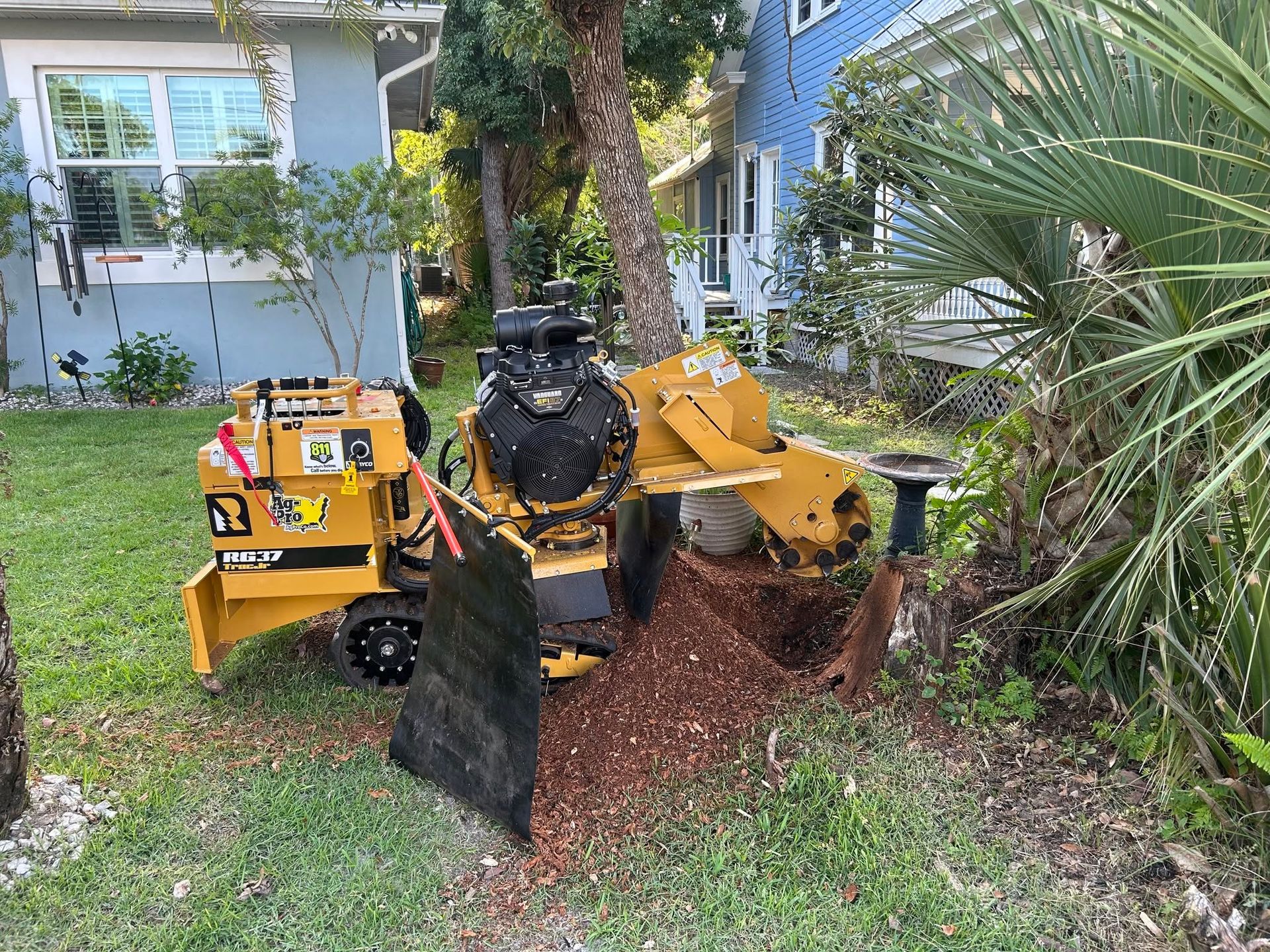 Yellow lawn tractor with front attachment parked on grass beside a house and palm tree.
