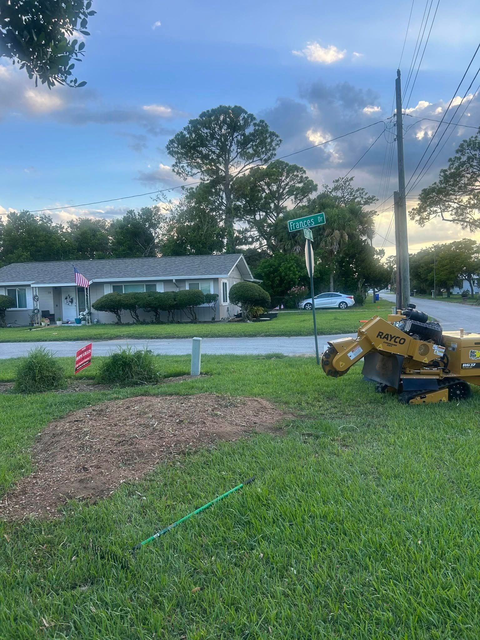 Yellow mini excavator beside a mulch pile on a grassy lawn at dusk, with a house and trees in the background