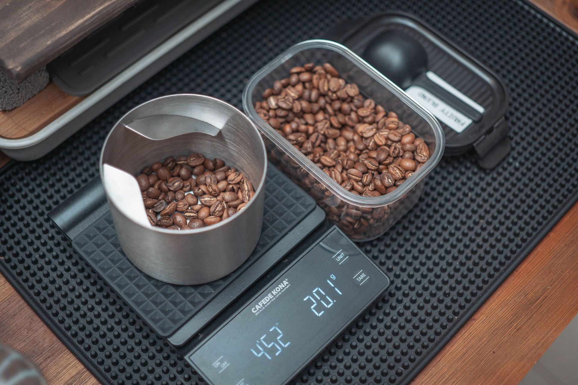 Weighing roasted coffee beans in a stainless steel cup on a digital scale beside a container of beans.
