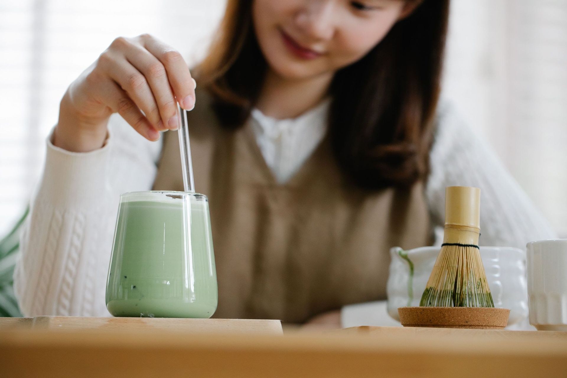 A woman stirring an iced matcha latte using a clear glass straw beside a traditional bamboo whisk on a wooden table.